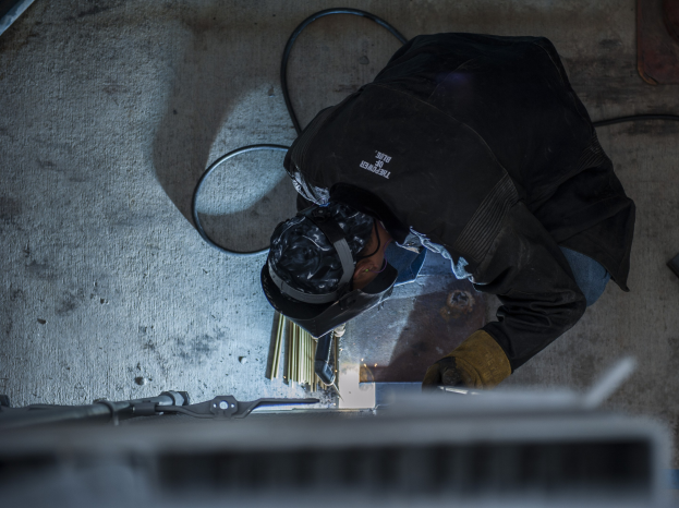 Ein Schweißer in Schutzausrüstung bedient eine Schweißmaschine an einem Metallstück, mit Werkzeugen und Gegenständen, die in der Nähe auf dem Boden verstreut sind.