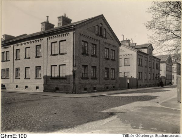 Ein Schwarz-Weiß-Foto einer Straßenszene mit Gebäuden, Straßenlaternen, Straßenlaternen, Kraftfahrzeugen, Bäumen und Himmel, mit dem Text "Tillhör Göteborgs Stads Museum" unten.