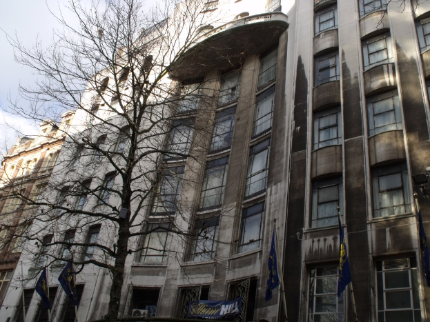 Gebäude mit Fenstern, einer Fahne und Flaggen an Stangen unten sowie einem Baum mit Ästen.