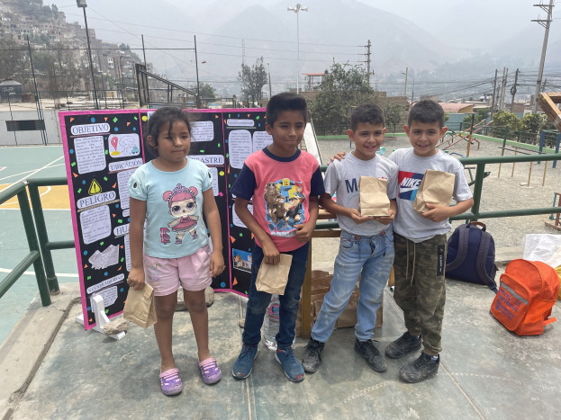 Eine Gruppe von Kindern auf einem Basketballfeld, die Papiertüten halten, mit Tüten und Gegenständen rechts und einer Tafel mit Text links, vor dem Hintergrund von Zäunen, Pfosten, Bäumen, Gebäuden, Hügeln und dem Himmel.