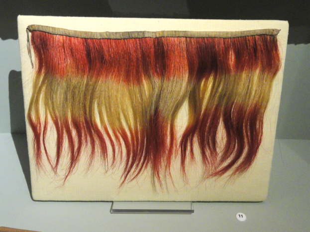 Nahaufnahme eines leuchtenden, bunt gemusterten Haarteil auf einem Brett in einer Museumsausstellung.