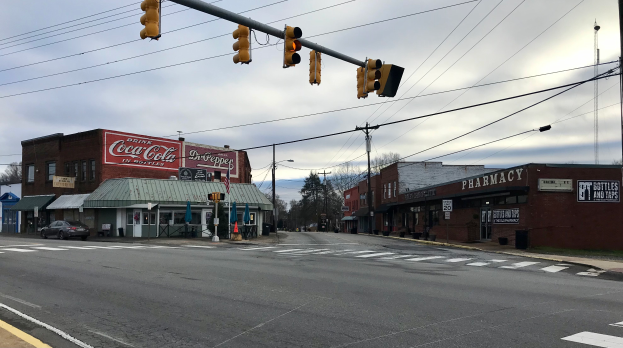 Eine Straßenkreuzung in einer Stadt mit Verkehrsampel, Fahrzeugen, Gebäuden mit Schildern, Strommasten mit Kabeln, Bäumen und einem bewölkten Himmel.