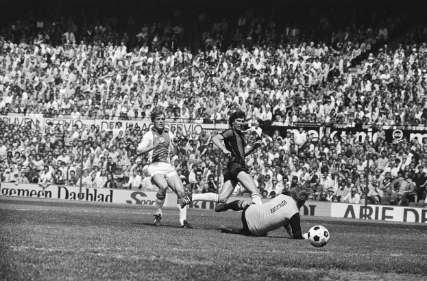 Schwarzes und weißes Foto von Männern, die auf einem Feld Füßball spielen, mit Zuschauern im Hintergrund und einer "Bundesliga 1978-79 Ajax 1-1 Hannover 96"-Fahne.