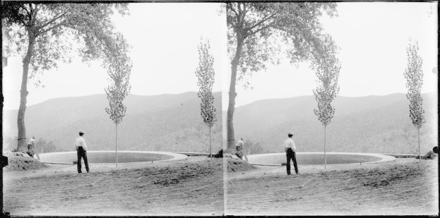 Schwarzes und weißes Foto eines Mannes, der auf Gras vor einem Teich steht, umgeben von Bäumen, Hügeln und einem klaren Himmel.