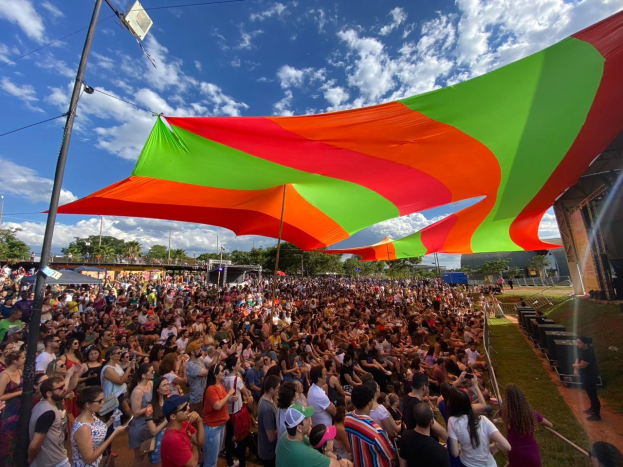 Eine große Menge steht vor einer Regenbogenflagge mit einem Gebäude, einer Bühne, Bäumen und Wolken im Hintergrund bei einer Festival-Jubiläumsfeier.