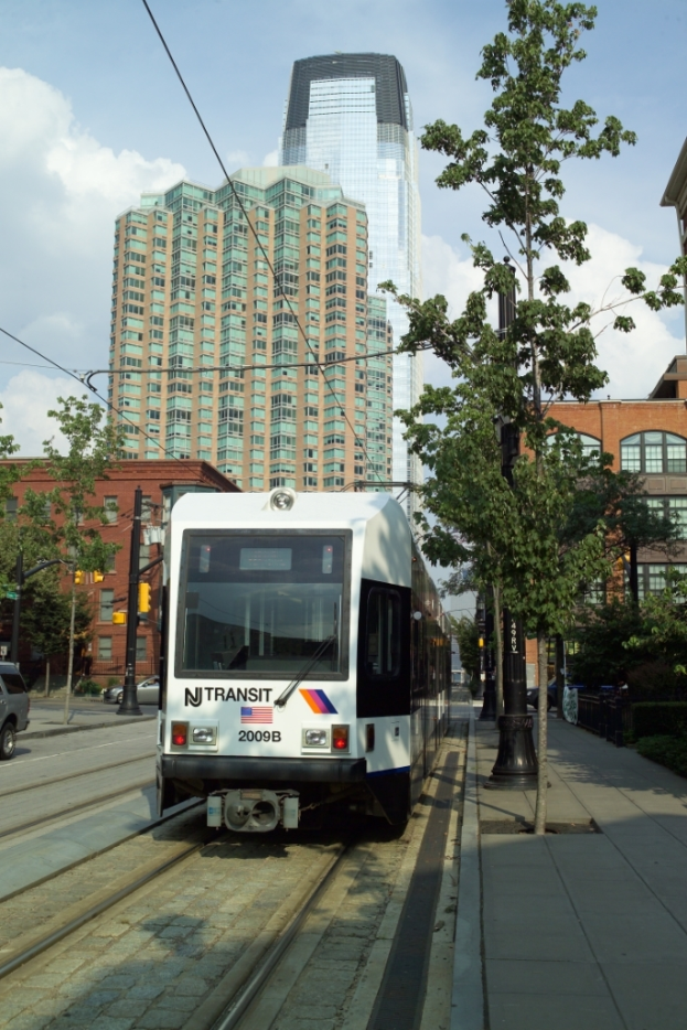 Ein Zug auf Schienen mit elektrischen Kabeln ├╝ber ihm, umgeben von B├Ąumen, Masten und Geb├Ąuden, mit einem Wolkenkratzer und Himmel im Hintergrund.