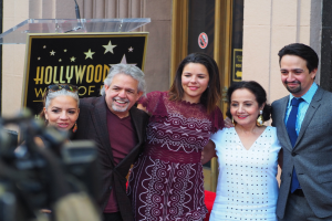 Eine Gruppe von Menschen posiert vor einem Hollywood Walk of Fame-Star mit einem Podium und einem Mikrofon, einer Tafel mit Text dahinter, einer Person mit einer Kamera links und einer Tür im Hintergrund.