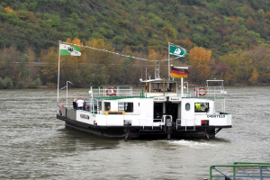 Ein Fährboot fährt einen Fluss hinunter, geschmückt mit Fahnen und Masten, mit ein paar Menschen an Bord, einem Geländer auf der rechten Seite und einem Hügel mit Bäumen im Hintergrund.