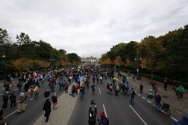 Eine große Gruppe von Menschen marschiert eine baumbestandene Straße mit Laternenpfählen entlang, hält Kameras in der Hand, mit einem Gebäude im Hintergrund und einem klaren Himmel.