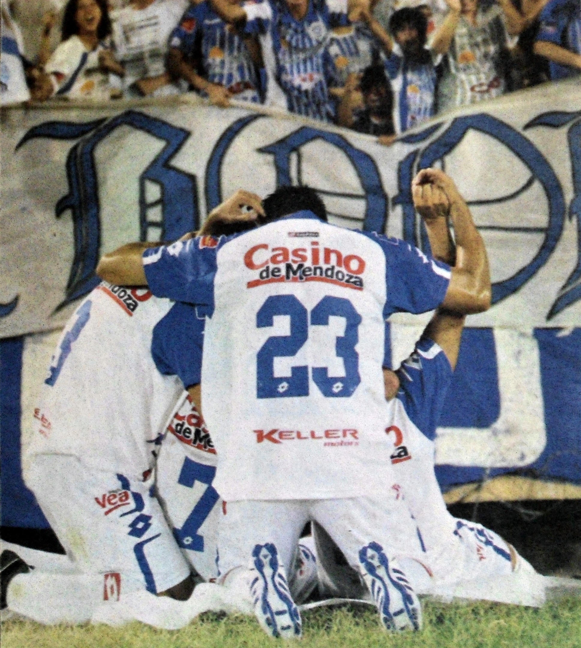 Eine Gruppe von Fußballspielern, die ein Tor feiern, mit einem Banner im Hintergrund und einer jubelnden Menge.