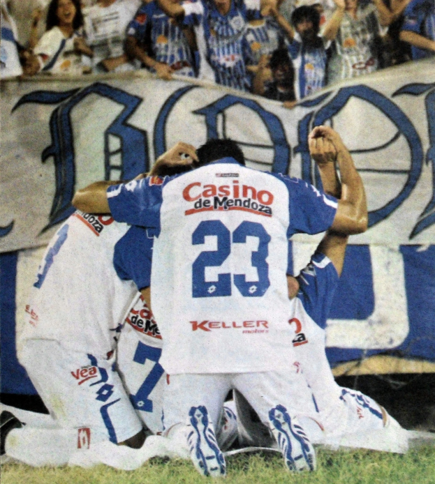 Eine Gruppe von Fußballspielern, die ein Tor feiern, mit einem Banner im Hintergrund und einer jubelnden Menge.