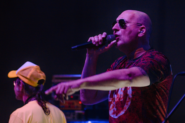 Ein Mann in rotem Hemd singt auf der Bühne in ein Mikrofon, neben ihm steht eine Person in einem gelben T-Shirt und einer gelben Mütze, vor dunklem Hintergrund.