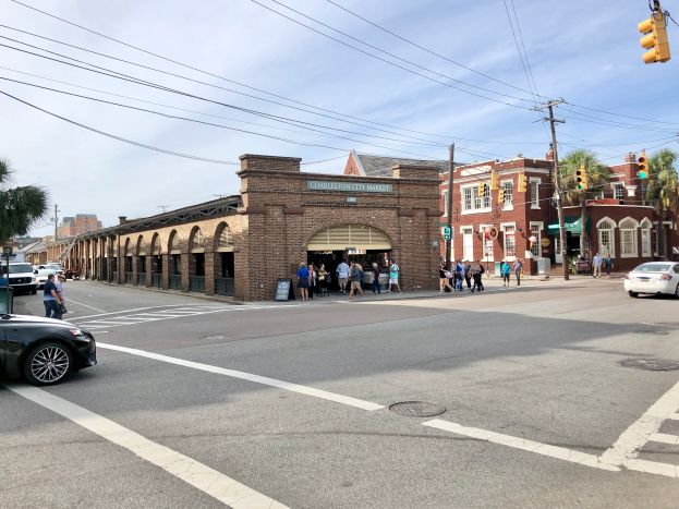 Eine Gruppe von Menschen überquert eine Straße in Charleston, South Carolina, in der Nähe des Charleston City Markets, mit Fahrzeugen, Gebäuden, Bäumen, Strommasten und einem bewölkten Himmel im Hintergrund.