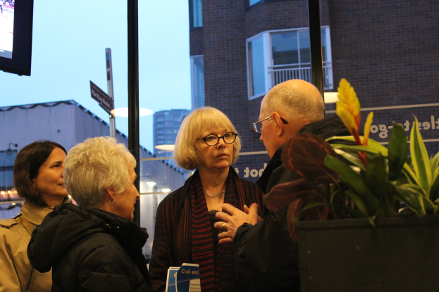 Eine Gruppe von Menschen, die vor einem Fernseher stehen und sprechen, mit einer Topfpflanze im Vordergrund und Gebäuden, Polen, Brettern, Lichtern und einem klaren blauen Himmel im Hintergrund.