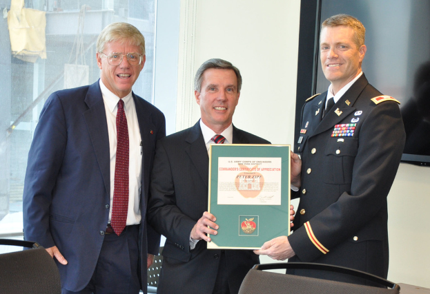 Drei Männer stehen, zwei halten ein gerahmtes Zertifikat, mit Stühlen, einem Fernseher und Glasfenstern im Hintergrund, die Baustoffe und einen Bau zeigen.