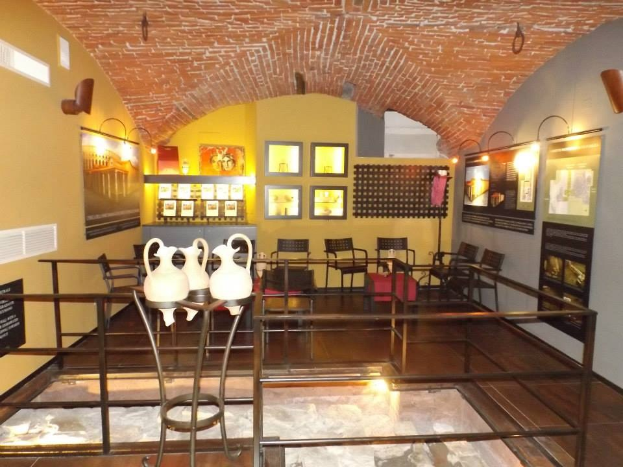 Ein Museumsraum mit mehreren Tischen, Stühlen, Displaytafeln, gerahmten Gegenständen an den Wänden, Vasen, Beleuchtung und anderen Gegenständen.