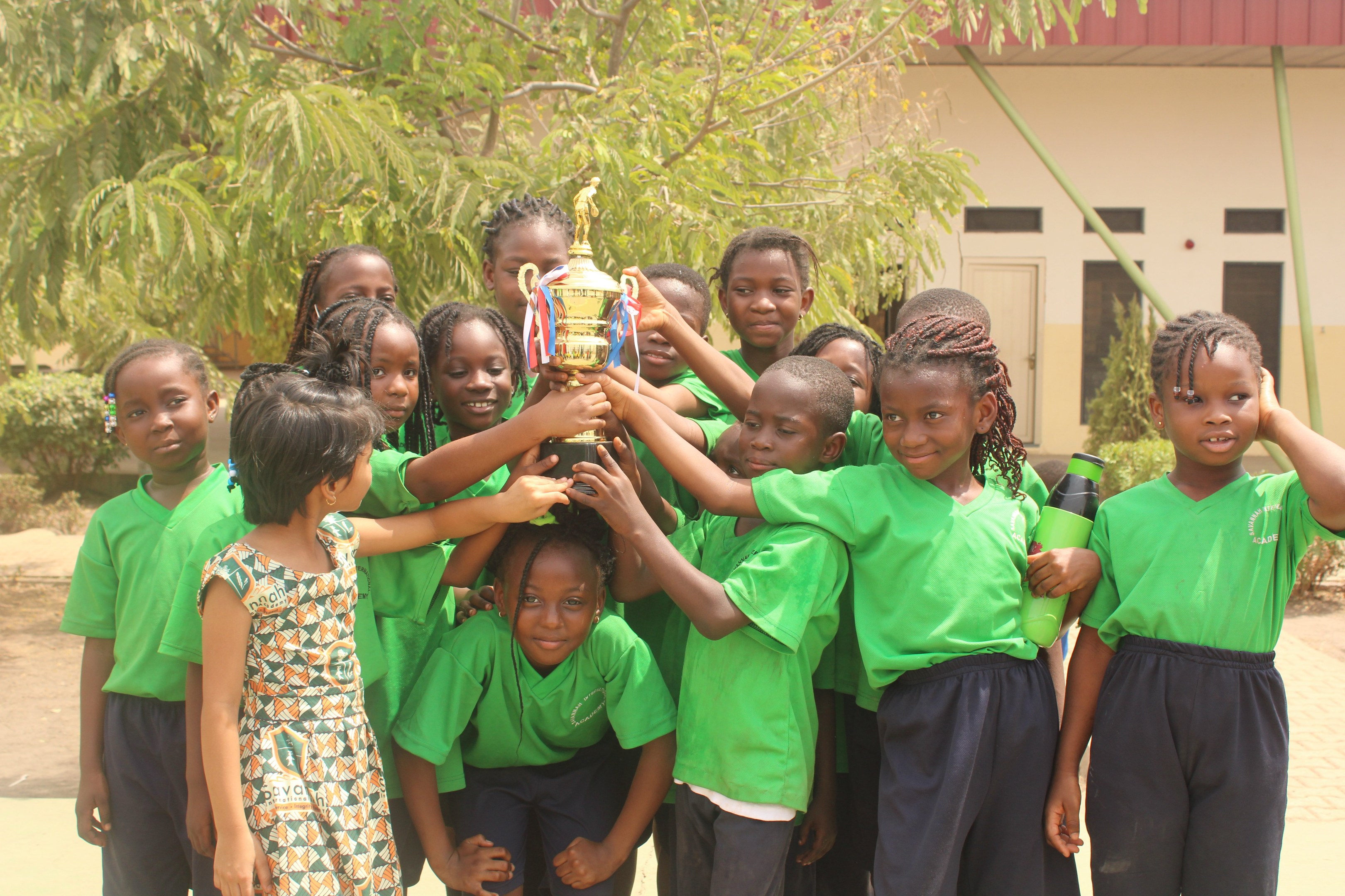 Eine Gruppe von Kindern hält eine Trophäe im Freien neben Bäumen und einem Gebäude.