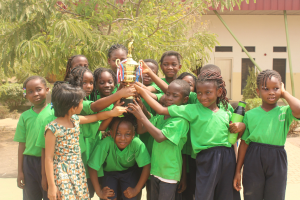 Eine Gruppe von Kindern hält eine Trophäe im Freien neben Bäumen und einem Gebäude.