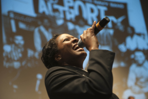 Eine Frau in einem schwarzen Kleid mit einem Dutt singt leidenschaftlich in ein Mikrofon auf der Bühne, ihr Gesicht von hellen Scheinwerfern beleuchtet, die Augen vor Emotion geschlossen.