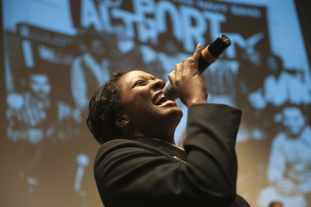 Eine Frau in einem schwarzen Kleid mit einem Dutt singt leidenschaftlich in ein Mikrofon auf der Bühne, ihr Gesicht von hellen Scheinwerfern beleuchtet, die Augen vor Emotion geschlossen.