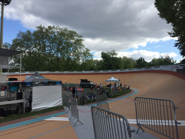 Gruppe von Radfahrern auf einer Schmutzspur bei den UCI BMX-Weltmeisterschaften 2018, mit Zelten, Fahrzeugen, Geländern, Bäumen und einem bewölkten Himmel im Hintergrund.