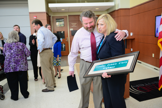Eine Gruppe von Menschen, die drinnen stehen, einer hält ein Foto mit 'Senator Debbie Clary Highway' darauf, mit einer Flagge, einer Wand, einer Tür und Lichtern im Hintergrund.