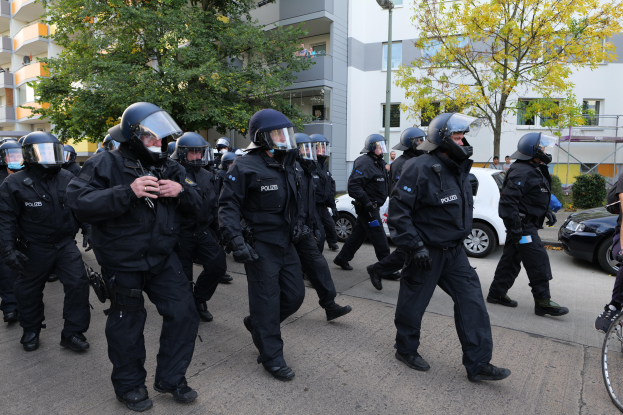 Eine Gruppe von Polizisten in Einsatzausrütstät marschiert auf einer Straße, mit einer Person, die ein Fahrrad hält, auf der rechten Seite, Fahrzeugen, Bäumen, Gebäuden und einem Laternenmast im Hintergrund.