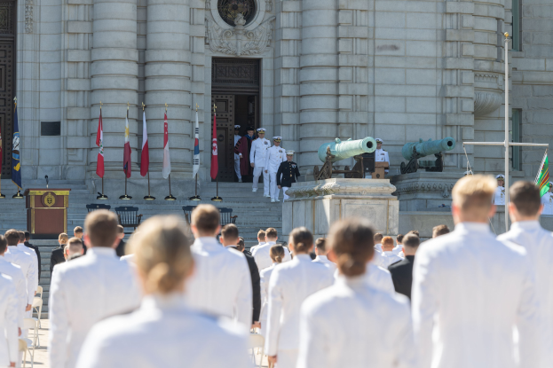 Gruppe von Menschen in weißen Marineuniformen auf einer Treppe vor einem Gebäude mit Säulen, die Fahnen halten und sich in der Nähe eines Podiums während einer Abschlussfeier versammeln.