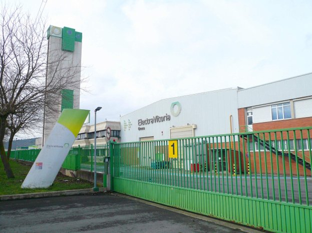 Ein großes Gebäude mit einem grünen Zaun davor, umgeben von einer Straße, Gras, Bäumen, Laternenmasten und einer Tafel, unter einem bewölkten Himmel, das als Hauptsitz von Electrovictoria dient.