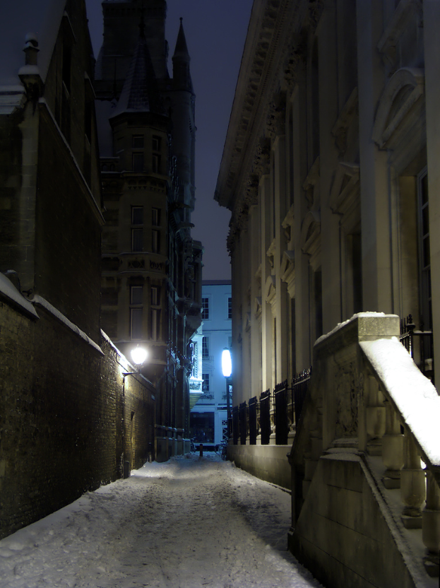 Eine schneebedeckte Straße mit Gebäuden und einem Geländer auf der rechten Seite, einer Wand auf der linken Seite und einem Himmel darüber.