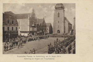 Ein Schwarz-Weiß-Foto eines Umzugs in Insterburg am 5. September 1914 mit vielen Menschen, Gebäuden und bewölktem Himmel sowie Text am unteren Rand.