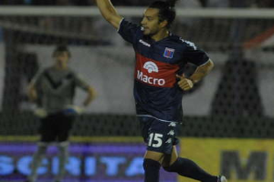 Ein Fußballspieler feiert nach einem Tor auf dem Boden, mit jubelnden Zuschauern und einer Person im Hintergrund, alles vor einem unscharfen, dynamischen Hintergrund.