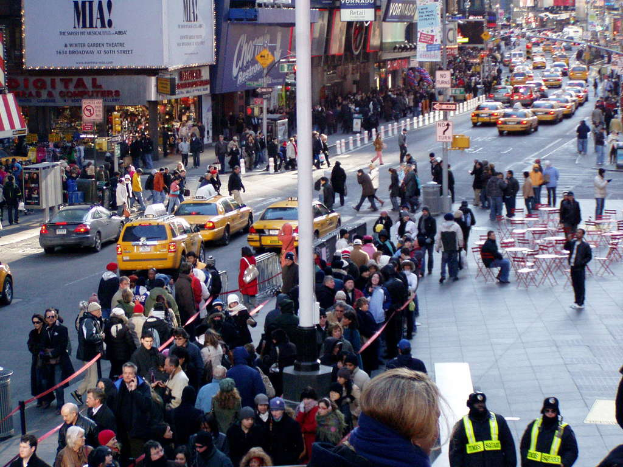 Fahrzeuge fahren auf einer Straße mit Fußgängern auf beiden Seiten, Gebäuden mit Geschäften und Namensschildern sowie verschiedener Straßenmöblierung wie Stühle, Tische, Laternen, Schilder und Ampeln.