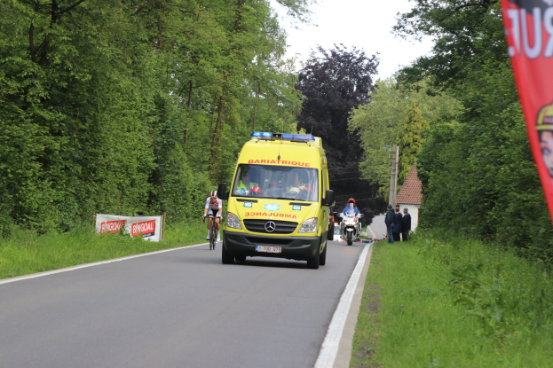 Ambulanz fährt auf einer Straße mit Fahrradfahrern daneben, gesäumt von Gras, Bäumen, Häusern, Strommasten und einem klaren blauen Himmel.
