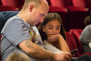 Zwei Menschen, ein erwachsener Mann und ein junges Mädchen, sitzen in einem Kino und sind beide auf ein Handy konzentriert, das der Mann hält.