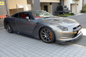 Ein silberner Nissan GT-R vor einem Gebäude mit Pflanzen und Gegenständen drumherum geparkt, und eine Tafel an der linken Seite der Gebäudewand befestigt.