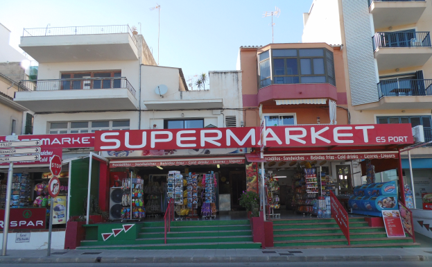 Ein Supermarkt an einer Straßenecke mit Gebäuden, Schildern und Pflanzen im Vordergrund und einem sichtbaren Himmel oben.