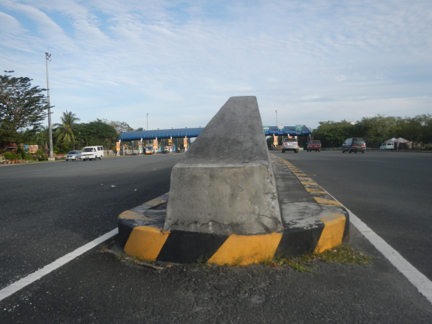 Ein Betonbollard an der Straße mit fahrenden Autos, Bäumen, Masten, Hütten und einem klaren blauen Himmel im Hintergrund.