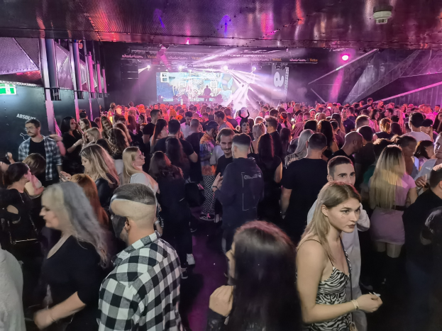 Eine große Gruppe von Menschen, die in einem Nachtclub stehen, beleuchtet von Deckenlampen und umgeben von Säulen, mit einem Bildschirm im Hintergrund, der auf ein Absolut Nightlife-Event hinweist.