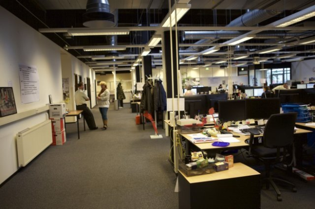 Ein geräumiges Büro mit zahlreichen Schreibtischen, Stühlen und Computergeräten, Menschen bei der Arbeit, Wände mit Tafeln geschmückt und große Fenster im Hintergrund.