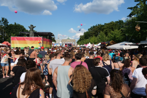 Eine große Menschenmenge, die eine Straße mit Zelten, Bäumen, Pfählen, Lichtern und einer Statue gesäumt, mit Gebäuden im Hintergrund und einem bewölkten Himmel mit Ballons, bei der Christopher Street Day Parade in Berlin.