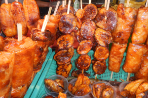 Ein Marktstand mit Spießen von Hähnchenflügeln, verschiedenen Lebensmitteln in Plastikbehältern und Menschen sowie anderen Gegenständen im Hintergrund.
