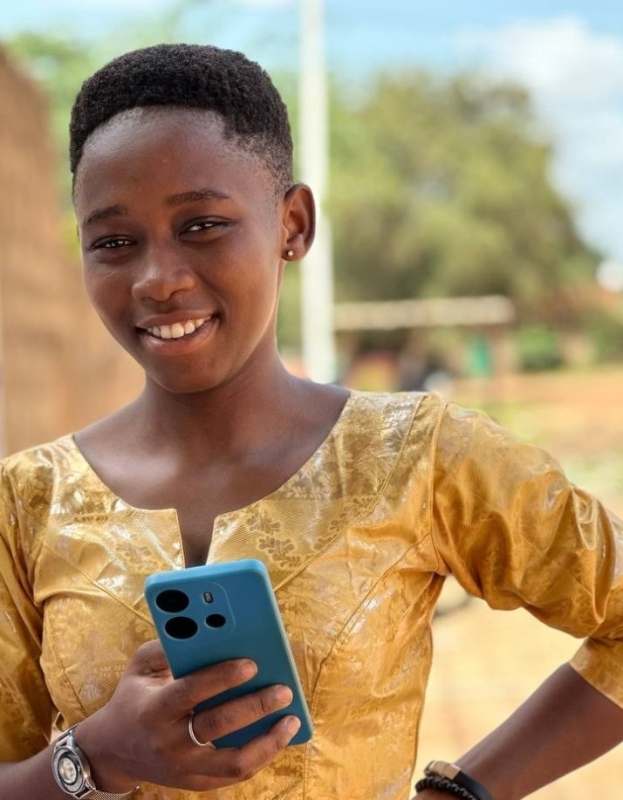 Ein junges Mädchen in einem gelben Kleid hält ein blaues Telefon, lächelt vor einer Wand, einem Pfahl, Bäumen und dem Himmel.