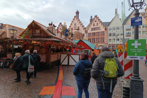 Eine Gruppe von Menschen, die über einen Kopfsteinpflasterweg neben einem Weihnachtsmarkt in Nürnberg, Deutschland, gehen, mit Laternenmästen und Informationsschildern in der Nähe und Gebäude im Hintergrund bei einem bewölktem Himmel.