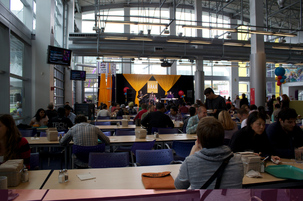 Eine Gruppe von Menschen an Tischen mit Flaschen, Taschentüchern und Tellern in einem Saal, mit einer gelben und schwarzen Bühne im Hintergrund, auf der Musiker spielen.