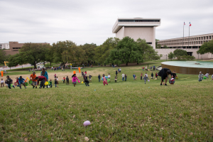 Menschen beim Ostereier-Spiel in einem grünen Feld mit Bäumen, Gebäuden, Fahnen und einem Brunnen im Hintergrund bei einem bewölkten Himmel.