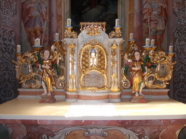 Ein Altar in einer Kirche mit Skulpturen von Menschen und Kerzen auf Ständern, vor einer Wand mit einem Gemälde, das eine feierliche Atmosphäre schafft.