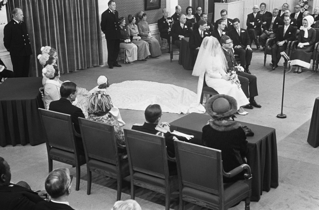 Schwarz-weiß-Foto einer Hochzeitszeremonie mit sitzenden und stehenden Gästen, einer Frau mit einem Blumenstrauß in der Mitte, einem Mikrofon auf einem Ständer rechts, Blumengestecken auf den Tischen links und Vorhängen mit gerahmten Fotos an der Wand im Hintergrund.