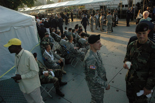 Eine Gruppe von Menschen in militärischer Uniform versammelt sich im Freien mit Zelten und einem Baum im Hintergrund, einige sitzen in der Mitte auf Stühlen.