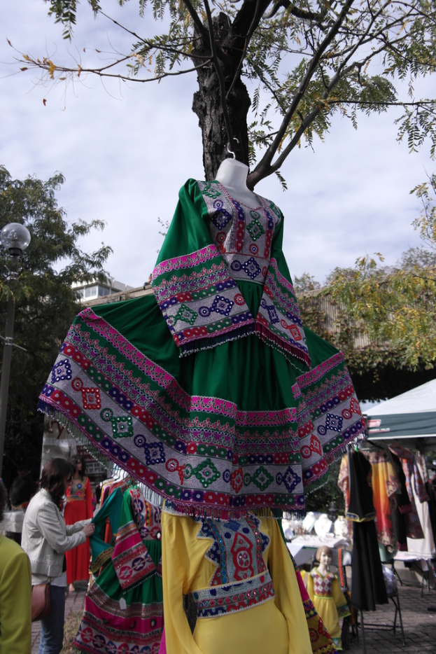 Eine Schaufensterpuppe steht inmitten eines belebten Einkaufsgebiets mit mehreren Geschäften, einer Person, die Kleider an einer Stange arrangiert, einem Gebäude und Bäumen im Hintergrund.